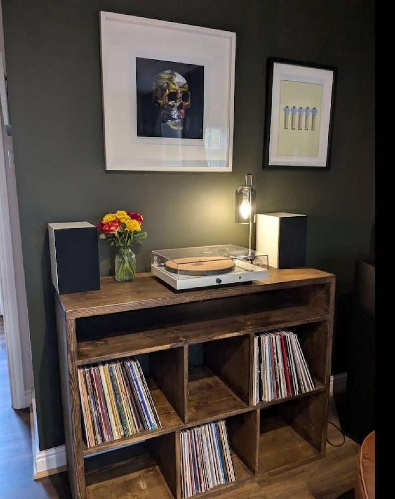 May include: A dark wood record player stand with six shelves, filled with vinyl records. A turntable sits on the top shelf, with a framed print of a skull above it.