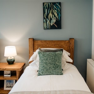 May include: A bedroom scene featuring two twin beds with wooden headboards. Each bed has white linens, a decorative pillow, and a gray throw. A nightstand with a lamp and a framed picture sits between the beds. Artwork hangs on the wall.