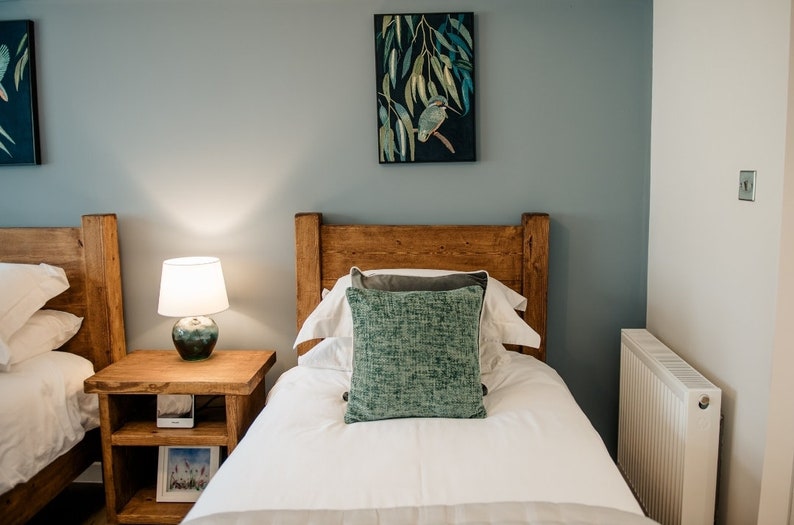 May include: A bedroom scene featuring two twin beds with wooden headboards. Each bed has white linens, a decorative pillow, and a gray throw. A nightstand with a lamp and a framed picture sits between the beds. Artwork hangs on the wall.