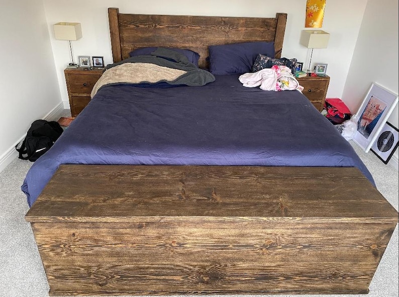 K&ouml;nnte beinhalten: Ein dunkelbraunes Holzbettgestell mit einer blauen Bettdecke und einem passenden Holz-Fu&szlig;teil mit Stauraum. Das Bett ist aus dunkelbraunem Holz gefertigt und hat ein schlichtes Design.