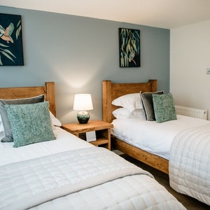 May include: A bedroom with two twin beds featuring wooden headboards. Each bed is made with white linens, gray and teal pillows, and a quilted bedspread. Artwork with birds hangs on the blue-gray wall. A lamp sits on a wooden nightstand.