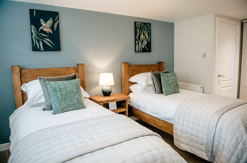 May include: A bedroom with two twin beds featuring wooden headboards. Each bed is made with white linens, gray and teal pillows, and a quilted bedspread. Artwork with birds hangs on the blue-gray wall. A lamp sits on a wooden nightstand.