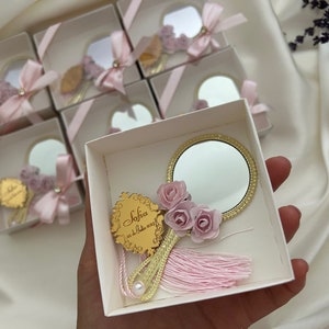 Puede incluir: Una peque&ntilde;a caja blanca con una cinta rosa y un espejo dorado con un borla rosa y flores. El espejo tiene un marco dorado y la caja tiene una inscripci&oacute;n dorada que dice "Sofia 02.12.2023".