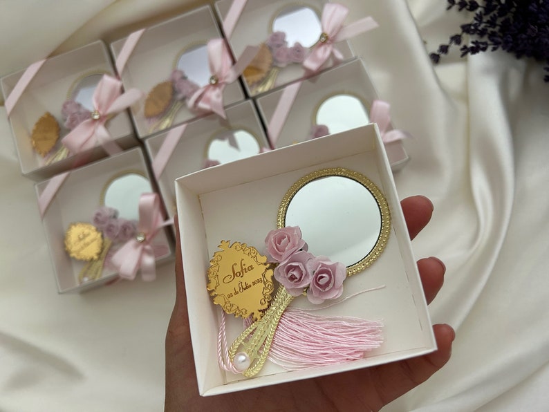 Puede incluir: Una peque&ntilde;a caja blanca con una cinta rosa y un espejo dorado con un borla rosa y flores. El espejo tiene un marco dorado y la caja tiene una inscripci&oacute;n dorada que dice "Sofia 02.12.2023".