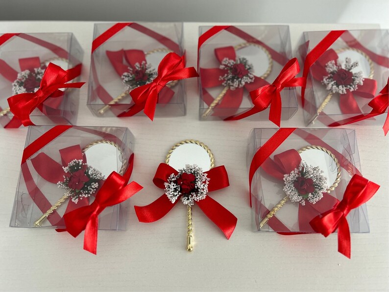 May include: A set of small, clear boxes with red ribbon bows, each containing a small, gold-handled mirror with a white and red flower embellishment.