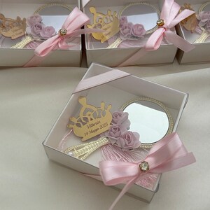 Puede incluir: Una peque&ntilde;a caja blanca con una cinta rosa y un peque&ntilde;o espejo en su interior. El espejo est&aacute; decorado con una corona dorada y flores rosas. La caja tiene una inscripci&oacute;n personalizada que dice "Vittoria 14 Maggio 2023".
