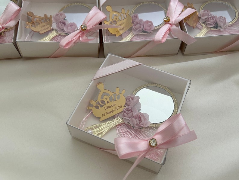 Puede incluir: Una peque&ntilde;a caja blanca con una cinta rosa y un peque&ntilde;o espejo en su interior. El espejo est&aacute; decorado con una corona dorada y flores rosas. La caja tiene una inscripci&oacute;n personalizada que dice "Vittoria 14 Maggio 2023".