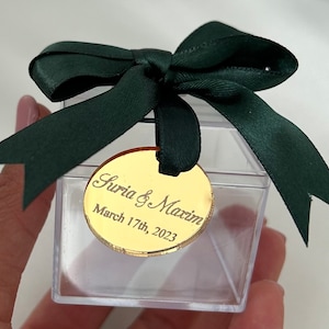 May include: A clear plastic box with a green satin ribbon tied in a bow. A gold round tag is attached to the ribbon with the text "Suria & Maxim March 17th, 2023".