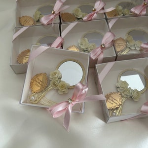 Puede incluir: Una colecci&oacute;n de peque&ntilde;as cajas blancas con lazos de cinta rosa, cada una contiene un peque&ntilde;o espejo redondo con marco dorado y un dise&ntilde;o floral. Las cajas est&aacute;n dispuestas en un patr&oacute;n escalonado sobre una superficie blanca.