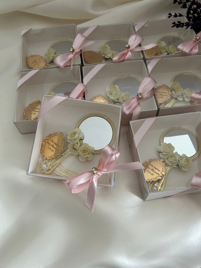 Puede incluir: Una colecci&oacute;n de peque&ntilde;as cajas blancas con lazos de cinta rosa, cada una contiene un peque&ntilde;o espejo redondo con marco dorado y un dise&ntilde;o floral. Las cajas est&aacute;n dispuestas en un patr&oacute;n escalonado sobre una superficie blanca.