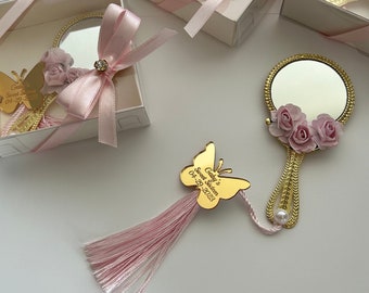 May include: A gold-colored hand mirror with a pink butterfly charm and a pink tassel. The mirror has a pink floral design and the butterfly charm has the text "Gabby's Sweet Sixteen 04-29-2023".