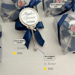 May include: A silver hand mirror with a blue ribbon and a white flower. The mirror has a personalized inscription that reads "Anjali's Quince 8/17/2024". The mirror is 4.13 inches in diameter. The mirror is available in silver or gold.