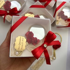 Puede incluir: Una peque&ntilde;a caja blanca con una cinta roja y un espejo dorado en su interior. El espejo tiene un dise&ntilde;o de rosa roja y una etiqueta dorada con el texto "Mia's Quinceanera January 21, 2023".