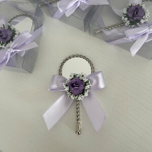 May include: A small, silver hand mirror with a purple flower and white flower accents. The mirror is tied with a lavender ribbon and bow.