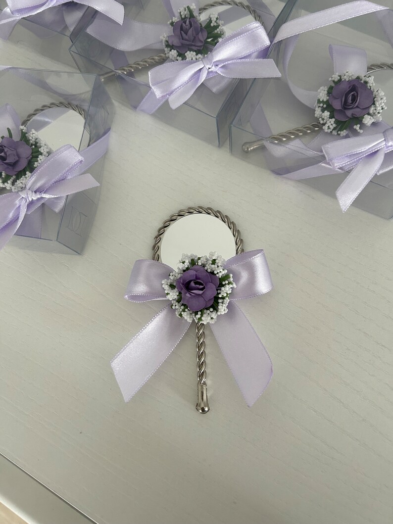 May include: A small, silver hand mirror with a purple flower and white flower accents. The mirror is tied with a lavender ribbon and bow.