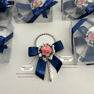May include: A small, silver hand mirror with a twisted handle, decorated with a navy blue ribbon bow and a pink flower. The mirror is packaged in a clear plastic box with a navy blue ribbon.