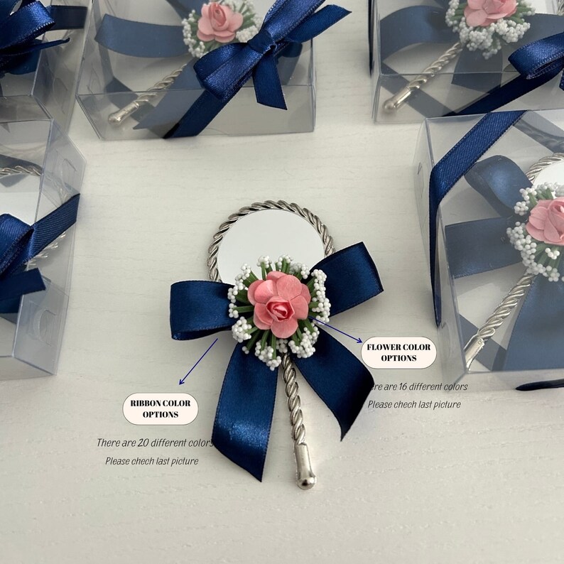 May include: A small, silver hand mirror with a twisted handle, decorated with a navy blue ribbon bow and a pink flower. The mirror is packaged in a clear plastic box with a navy blue ribbon.