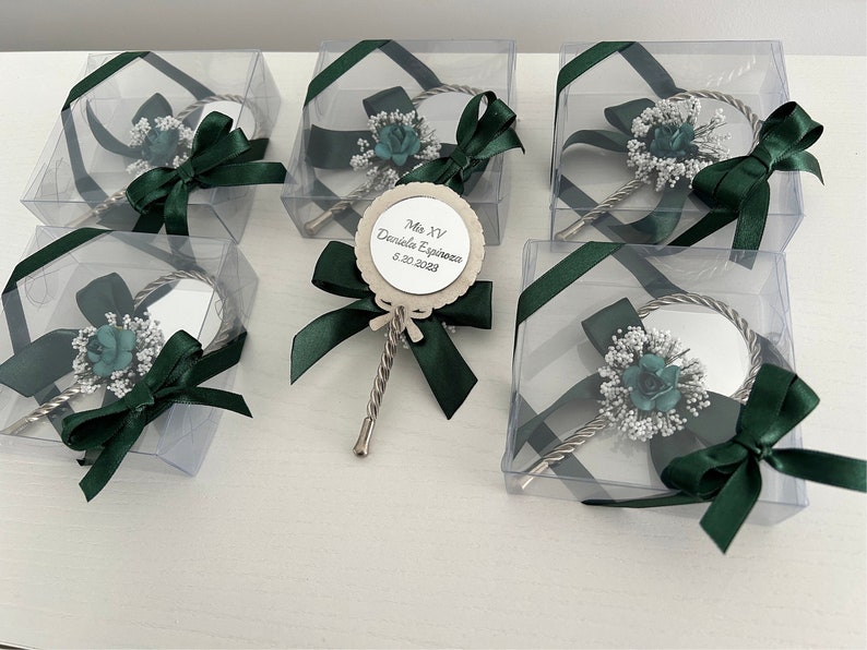 May include: A set of five clear plastic boxes with green ribbon bows, each containing a silver hand mirror with a small green flower and white flower embellishment. One box has a wooden circle with the text "Mis XV Daniela Espinoza 8.30.2023".