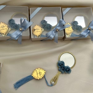 Puede incluir: Un conjunto de peque&ntilde;as cajas de regalo blancas con lazos de cinta azul. Cada caja contiene un peque&ntilde;o espejo redondo con marco dorado y un adorno de flor azul. Las cajas est&aacute;n dispuestas en fila sobre una superficie blanca. El texto "Sofia" es visible en el espejo.