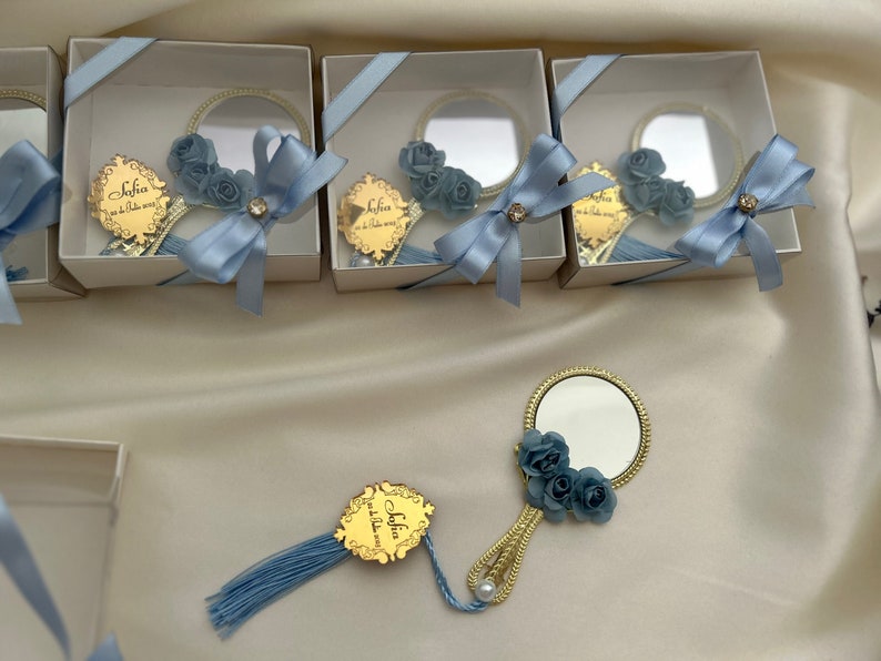 Puede incluir: Un conjunto de peque&ntilde;as cajas de regalo blancas con lazos de cinta azul. Cada caja contiene un peque&ntilde;o espejo redondo con marco dorado y un adorno de flor azul. Las cajas est&aacute;n dispuestas en fila sobre una superficie blanca. El texto "Sofia" es visible en el espejo.