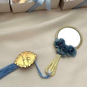 Puede incluir: Espejo de mano dorado con un borla azul y un dise&ntilde;o floral azul. El espejo tiene una peque&ntilde;a perla en el mango. El espejo est&aacute; unido a una etiqueta dorada con el texto "Sofia, &iexcl;eres la mejor!"