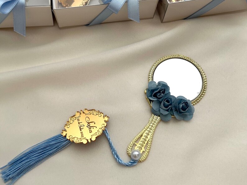 Puede incluir: Espejo de mano dorado con un borla azul y un dise&ntilde;o floral azul. El espejo tiene una peque&ntilde;a perla en el mango. El espejo est&aacute; unido a una etiqueta dorada con el texto "Sofia, &iexcl;eres la mejor!"