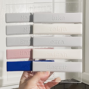 Peut inclure: Un ensemble de bandes rectangulaires en silicone fix&eacute;es sur une plaque en acrylique transparent. Les bandes sont grises, gris clair, roses et bleues, chacune portant des noms en relief : Mason, Luke, Mila, Reign, Emma, Ezra, Harper, Micah, Logan et Berrit.