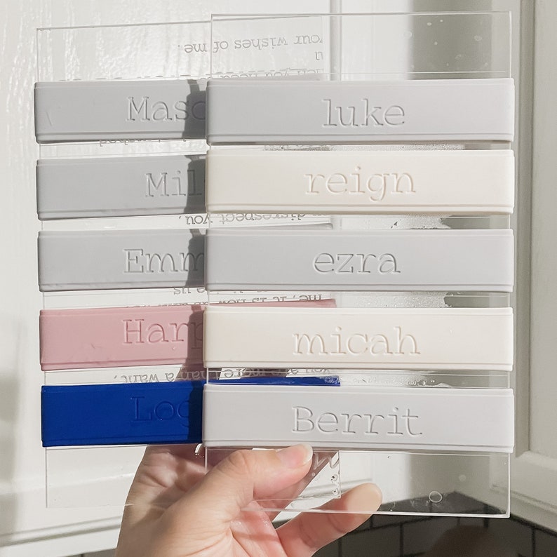 Peut inclure: Un ensemble de bandes rectangulaires en silicone fix&eacute;es sur une plaque en acrylique transparent. Les bandes sont grises, gris clair, roses et bleues, chacune portant des noms en relief : Mason, Luke, Mila, Reign, Emma, Ezra, Harper, Micah, Logan et Berrit.