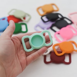 May include: A light green silicone slider buckle with a round opening in the center. The buckle is held in a hand, and there are other colorful silicone slider buckles in the background.