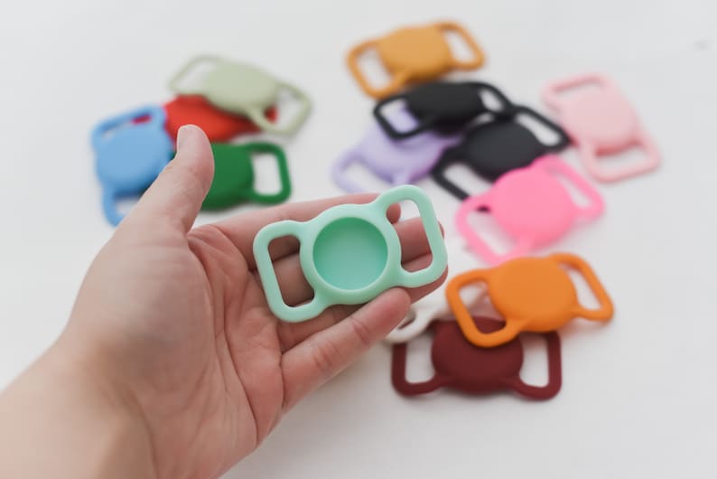 May include: A light green silicone slider buckle with a round opening in the center. The buckle is held in a hand, and there are other colorful silicone slider buckles in the background.