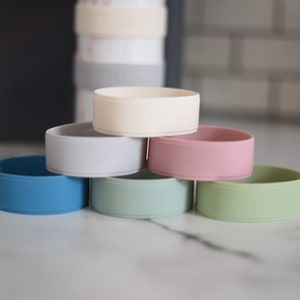Peut inclure: Un ensemble de six protections de bouteille circulaires en silicone de diff&eacute;rentes couleurs, dont cr&egrave;me, gris clair, rose, bleu, vert sauge et vert clair, empil&eacute;es en pyramide avec une bouteille plus haute visible en arri&egrave;re-plan flou.