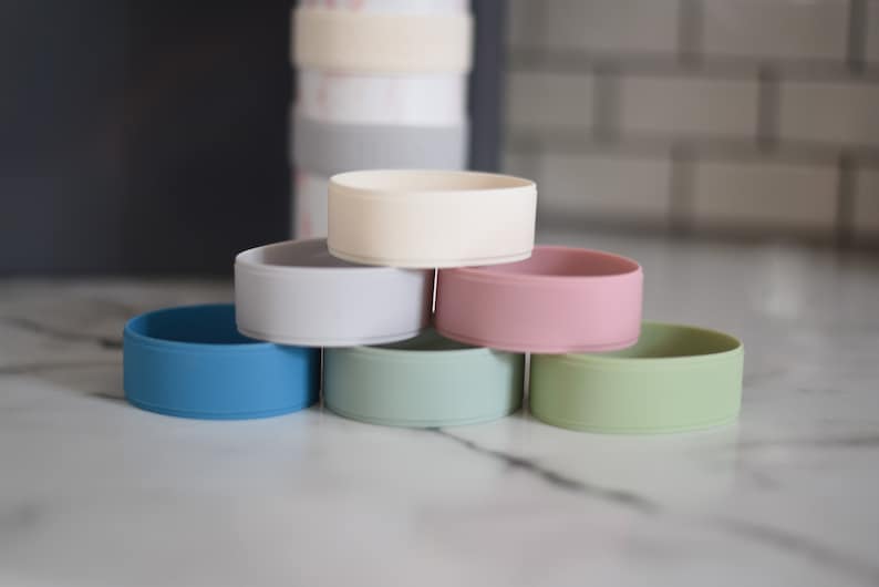 Peut inclure: Un ensemble de six protections de bouteille circulaires en silicone de diff&eacute;rentes couleurs, dont cr&egrave;me, gris clair, rose, bleu, vert sauge et vert clair, empil&eacute;es en pyramide avec une bouteille plus haute visible en arri&egrave;re-plan flou.