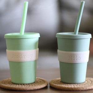 Peut inclure: Deux gobelets en silicone vert clair avec couvercles et pailles assortis. Chaque gobelet comporte une bande en silicone blanc au centre. Le gobelet de gauche porte le nom Audrey en relief, et celui de droite porte le nom Milo sur la bande blanche.