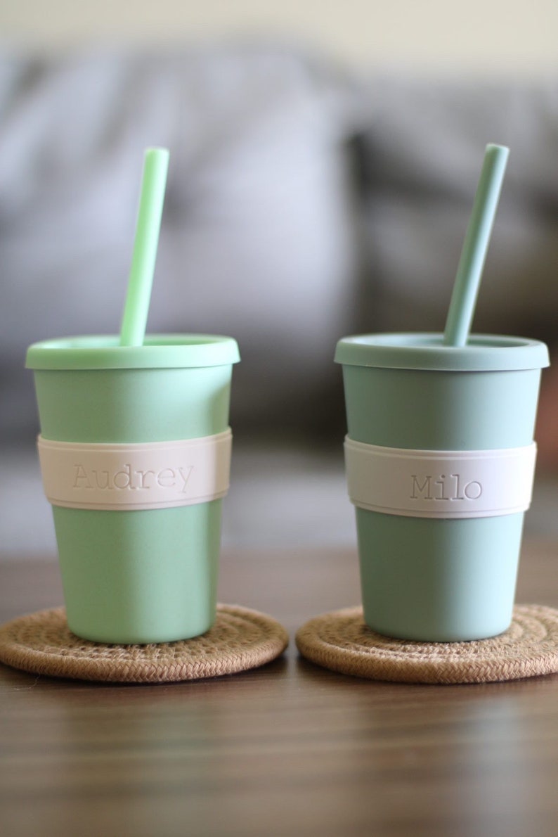 Peut inclure: Deux gobelets en silicone vert clair avec couvercles et pailles assortis. Chaque gobelet comporte une bande en silicone blanc au centre. Le gobelet de gauche porte le nom Audrey en relief, et celui de droite porte le nom Milo sur la bande blanche.