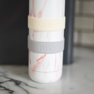Peut inclure: Une bouteille cylindrique blanche avec un motif de marbre rose comportant deux bandes en silicone. La bande sup&eacute;rieure est de couleur cr&egrave;me avec le nom Audrey en relief, et la bande inf&eacute;rieure est gris clair avec le nom Milo en relief.