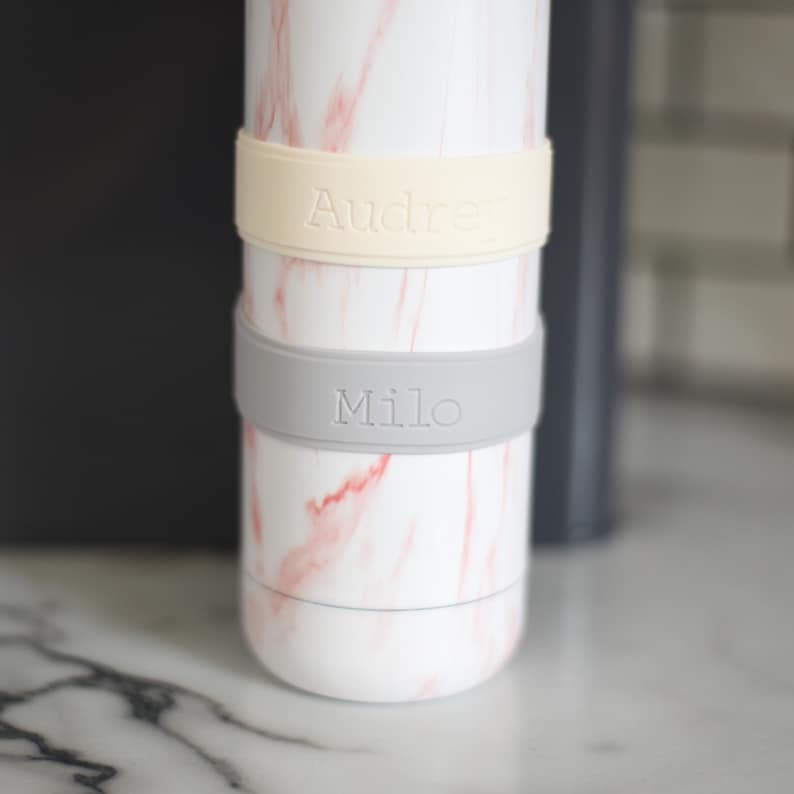 Peut inclure: Une bouteille cylindrique blanche avec un motif de marbre rose comportant deux bandes en silicone. La bande sup&eacute;rieure est de couleur cr&egrave;me avec le nom Audrey en relief, et la bande inf&eacute;rieure est gris clair avec le nom Milo en relief.