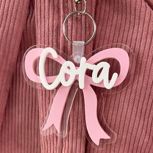 May include: A clear acrylic keyring shaped like a bow with a light pink bow overlay and the name Cora written in white script lettering across the centre. The keyring includes a silver metal ring attached by a small clear plastic loop.