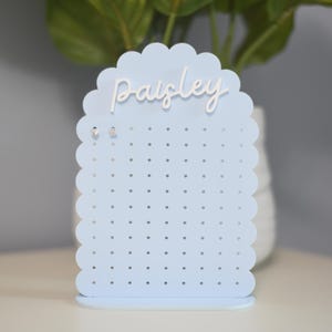 May include: A light blue earring display stand with a scalloped top edge and numerous small holes for holding earrings. The word "Paisley" is written in white script across the top. Two pairs of earrings are displayed.