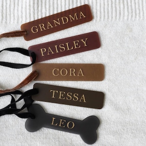 Puede incluir: Una colección de etiquetas personalizadas en varias formas y colores. Las etiquetas incluyen diseños rectangulares y en forma de hueso, con nombres como "GRANDMA", "PAISLEY", "CORA", "TESSA" y "LEO" en letras doradas. Cada etiqueta tiene una cinta o cordón.