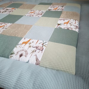 Könnte beinhalten: Eine Patchwork-Babydecke mit hellblauem Rand. Die Decke besteht aus verschiedenen Stoffen mit verschiedenen Tierdrucken, darunter Giraffen und Elefanten. Die Decke ist perfekt für ein Babyzimmer oder ein Spielzimmer.