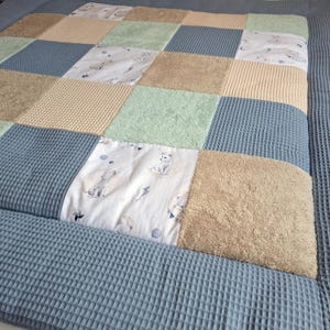 Könnte beinhalten: Eine Patchwork-Steppdecke mit Quadraten aus verschiedenen Texturen und Farben, darunter blaues Waffelgewebe, beigefarbener Frottee und hellgrün. Einige Quadrate haben einen weißen Stoff mit einem Cartoon-Wolf- und Mond-Design. Die Steppdecke hat einen blauen Waffelgewebe-Rand.