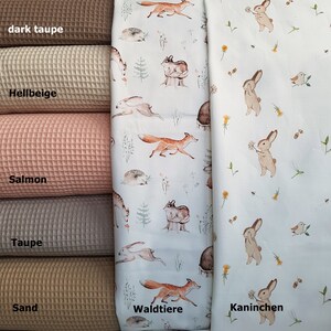 May include: Five different colors of waffle fabric swatches, from dark taupe to sand. The fabric is displayed next to two fabric swatches with different animal prints. One fabric features a white background with brown foxes, deer, and hedgehogs. The other fabric features a white background with brown rabbits and the text "Kaninchen". The text "Waldtiere" is printed on the fabric with the foxes, deer, and hedgehogs.