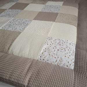 Könnte beinhalten: Eine beige und weiße Waffelstruktur-Babydecke mit Patchwork-Design. Die Decke besteht aus Quadraten mit unterschiedlichen Texturen und Mustern, darunter ein Blumendruck.