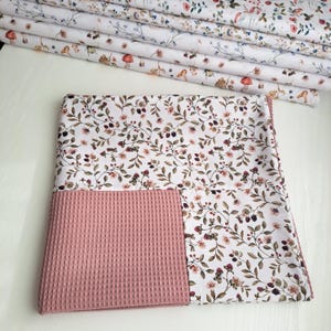 Könnte beinhalten: Ein gefalteter weißer Stoff mit einem floralen Muster aus rosa und lila Beeren und grünen Blättern. Ein Abschnitt aus rosa Waffelstoff ist unten angenäht. Im Hintergrund sind mehrere Rollen passenden Stoffes gestapelt.
