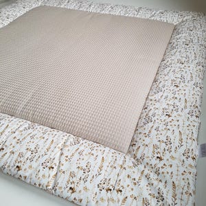 Könnte beinhalten: Eine beige Spielmatte aus Waffelgewebe mit einem weißen Rand, der mit einem floralen Muster aus braunen und goldenen Wildblumen verziert ist.