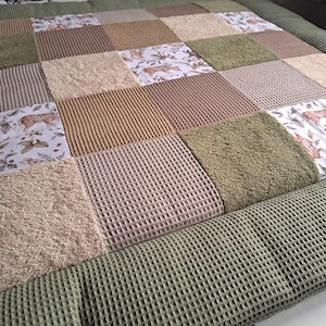 Könnte beinhalten: Eine Patchwork-Decke mit abwechselnden Quadraten aus olivgrünem Waffelstoff, beigem Frottee mit Struktur und einem weißen Baumwollstoff mit Waldmotiven wie Hirschen, Füchsen und Blättern. Die Decke hat einen breiten Rand aus olivgrünem Waffelstoff.