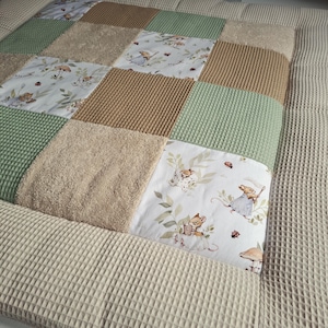 Könnte beinhalten: Eine Patchwork-Decke mit abwechselnden Quadraten aus salbeigrünem Waffelstoff, beigem Frottee und einem weißen Baumwollstoff mit Illustrationen von kleinen Mäusen, Pilzen und Marienkäfern, eingefasst mit einer beigen Waffelstoff-Bordüre.
