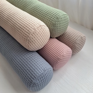 Könnte beinhalten: Eine Ansammlung zylindrischer Kissen in Pastelltönen wie Hellblau, Rosa, Beige und Grün. Jedes Kissen hat ein strukturiertes Waffelwebmuster.