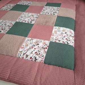 Könnte beinhalten: Ein Patchwork-Quilt mit Quadraten aus verschiedenen Texturen und Mustern. Der Quilt enthält Quadrate aus rosa Waffelstoff, grünem Waffelstoff, floralem Stoff mit Hasen und weichem beigefarbenem Stoff. Der Rand ist einfarbiger rosa Waffelstoff.