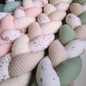 Könnte beinhalten: Ein geflochtener Baby-Bettumrandung in verschiedenen Farben und Texturen. Der Bumper hat Abschnitte aus rosa, cremefarbenem und grünem Waffelstoff sowie Abschnitte mit floralen und botanischen Drucken. Das Design kombiniert weiche und strukturierte Materialien.